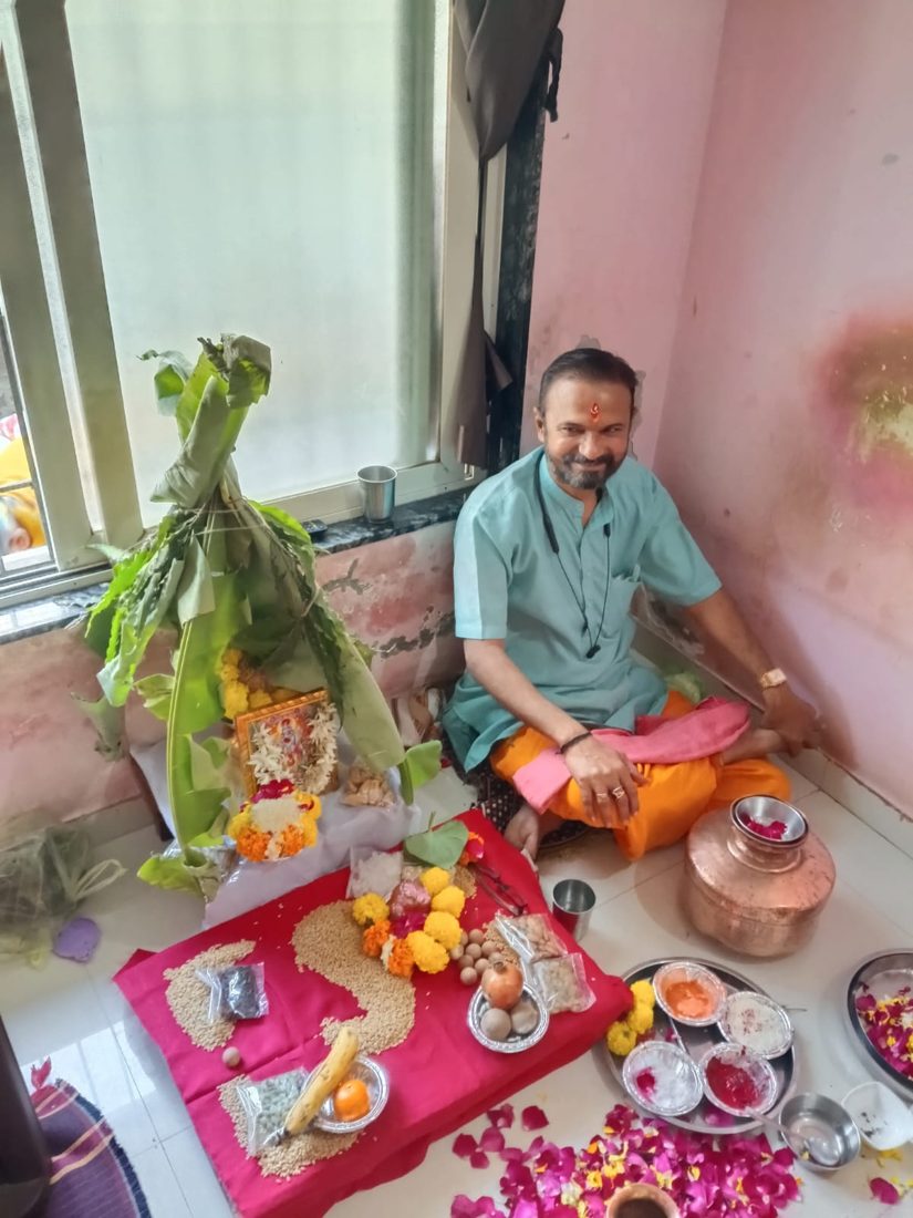 Priest seated beside a puja arrangement with a banana-leaf mandap, flowers and a copper kalash