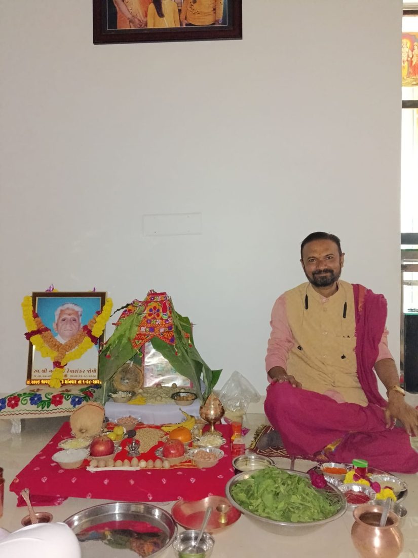 Priest seated beside a home puja arrangement with flowers, fruits and copper vessels