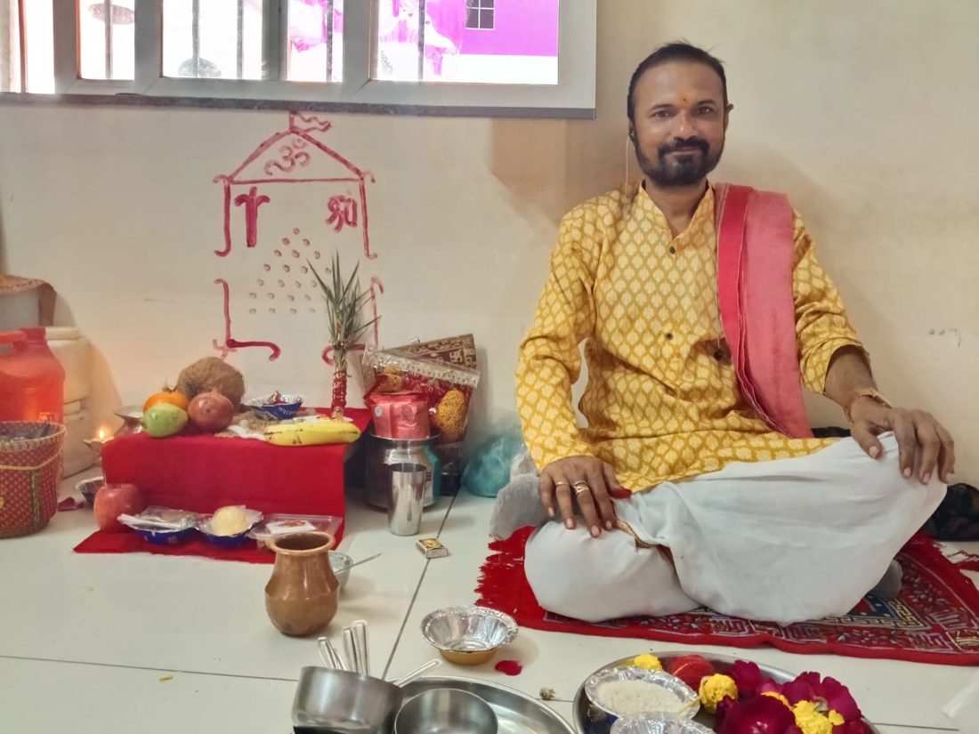 Priest seated before a puja setup with a hand-drawn Om and swastik on the wall